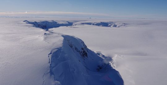 Loftmynd af snævi þöktum jökli með djúpum sigdölum og sprungum. Í forgrunni sést brött, dökk klettabrún sem sker sig í gegnum hvítan jökulflötinn. Landslagið er víðáttumikið og flatt í kring.
